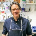 Headshot of Michael Velliquette wearing an artist's apron in a studio with works-in-progress in the background