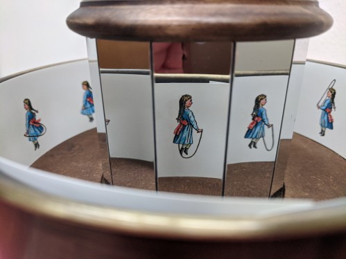 Zoetrope with a sequence of drawings of a girl with two braids wearing a blue dress with a large red sash jumping rope