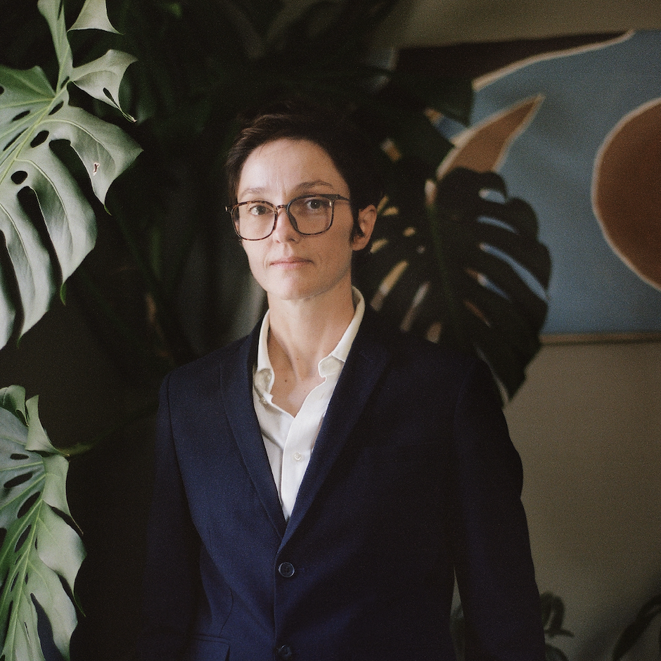 Headshot of M. Hamilton (they/them) in an interior room with a window-lit monstera plant on their right and an abstract painting on the wall in the background