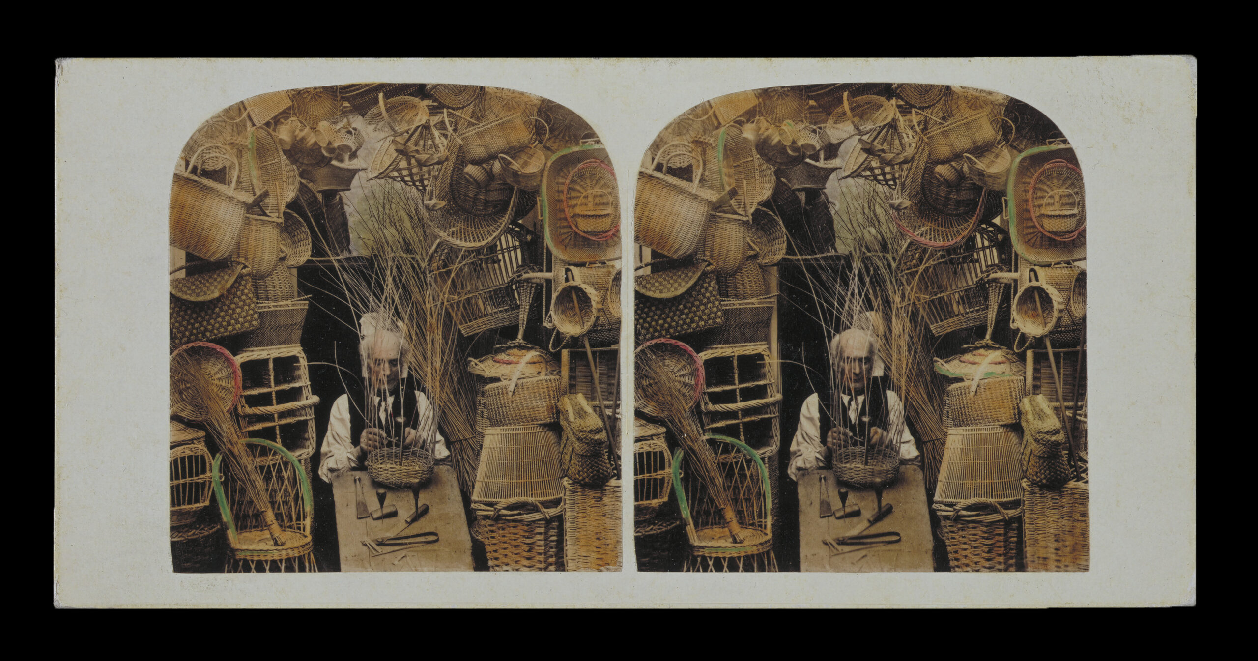 Steareoscope images, side by side, of a person with a bald head and white muttonchops, sitting among a towering wall of woven baskets. He is also weaving a basket. Most of the scene is sepia tones, but some of the basket rims have been hand-painted, red for some and green for others.